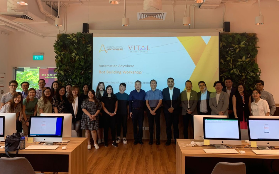 Group of about 25 people posing in a room with a projected slide reading "Automation Anywhere Bot Building Workshop."